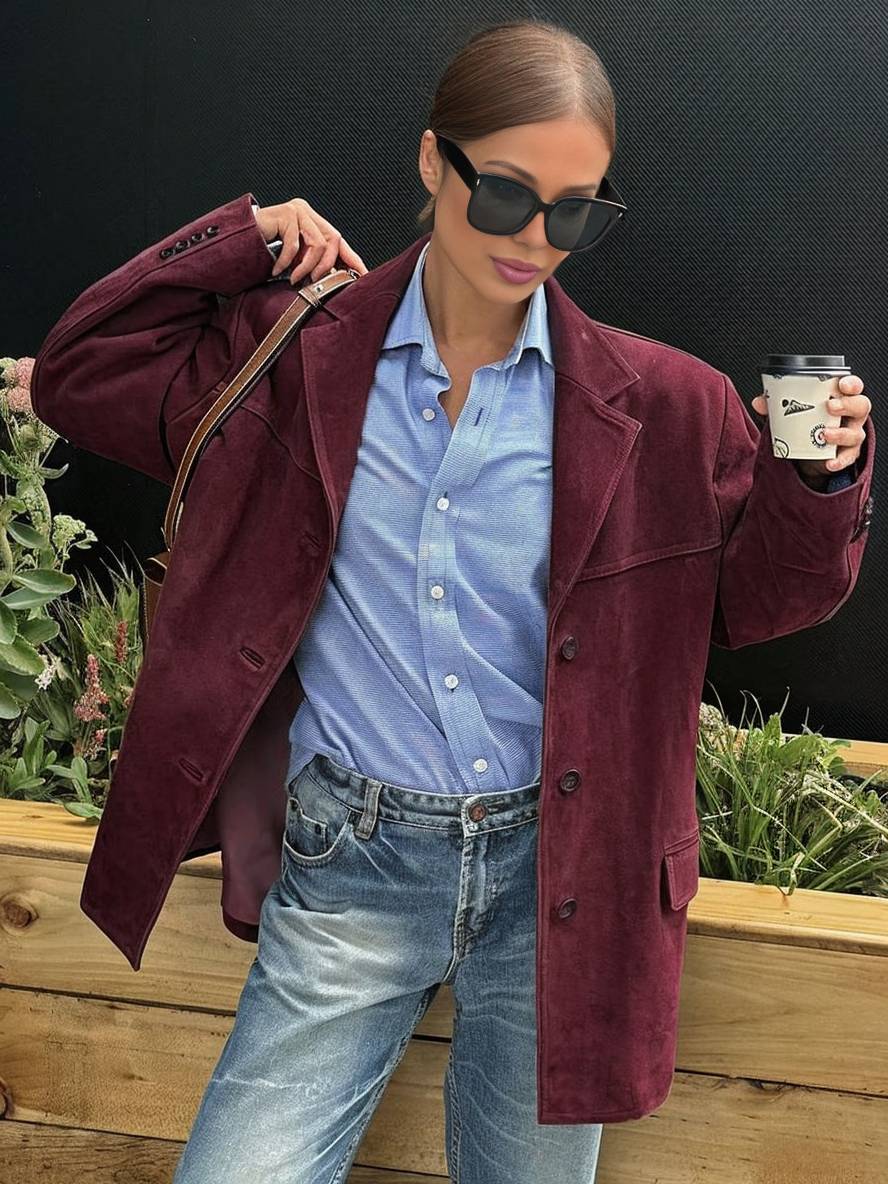 Donna con giacca in pelle marrone, camicia a righe blu, maglione marrone, jeans chiari, tiene caffè. Moda autunnale casual elegante.
