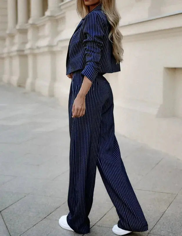 Donna in completo pantalone blu a righe, giacca corta, pantaloni larghi, scarpe bianche, sfondo urbano. Moda elegante, abbigliamento femminile.